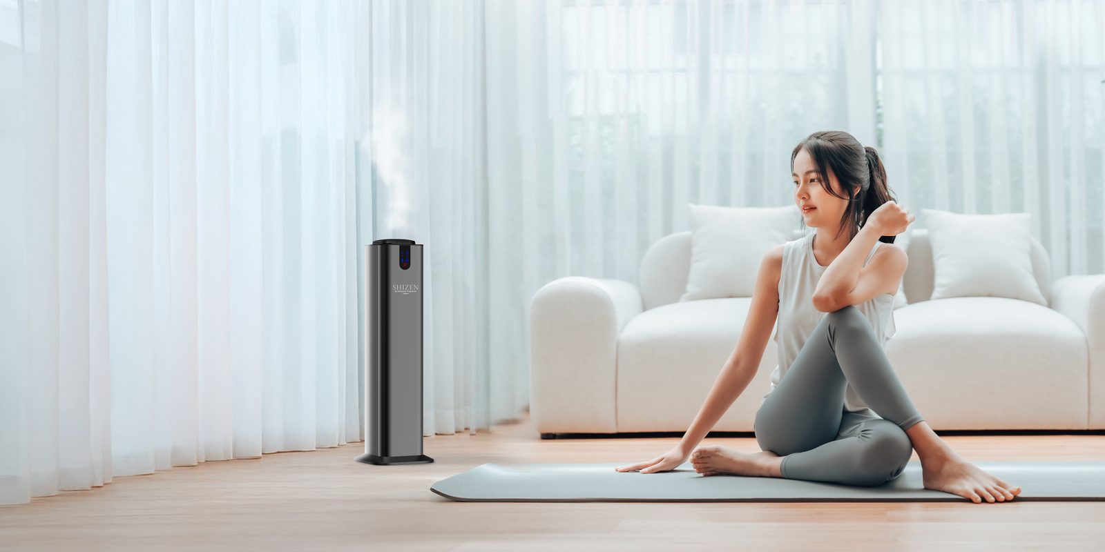 Ruang keluarga terang dengan tirai tipis — diffuser tower Shizen mengeluarkan kabut halus sementara seorang perempuan beristirahat di matras yoganya